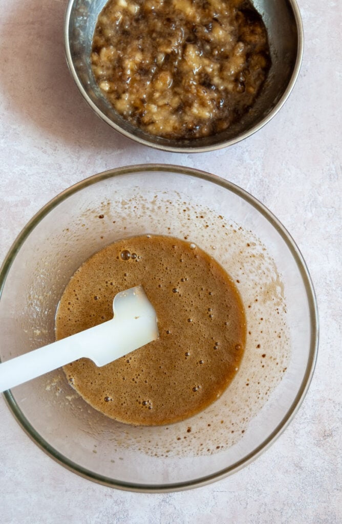 a silver bowl of overripe mashed bananas and a glass bowl of whisked brown sugar and eggs with a wooden spatula.