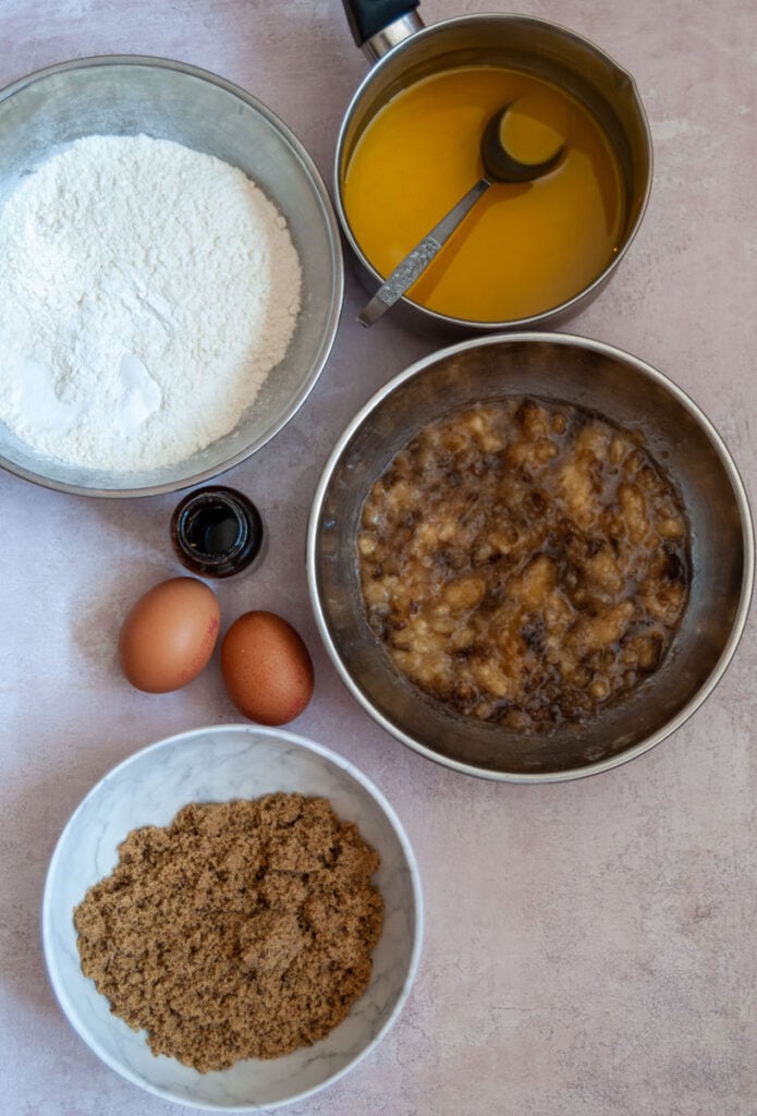 a white bowl of brown sugar, a silver bowl of flour, 2 eggs, a bowl of mashed banana and a pan of melted butter with a spoon