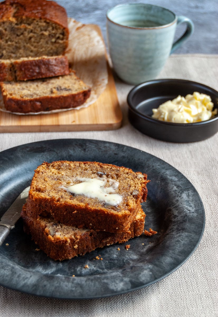 two slices of banana bread spead with butter on a black plate and large banana bread sitting on a wooden board with a blue cup of coffee and a black pot of butter.