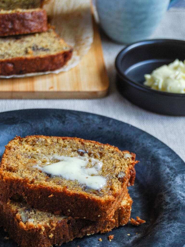 two slices of banana loaf spread with butter on a black plate.