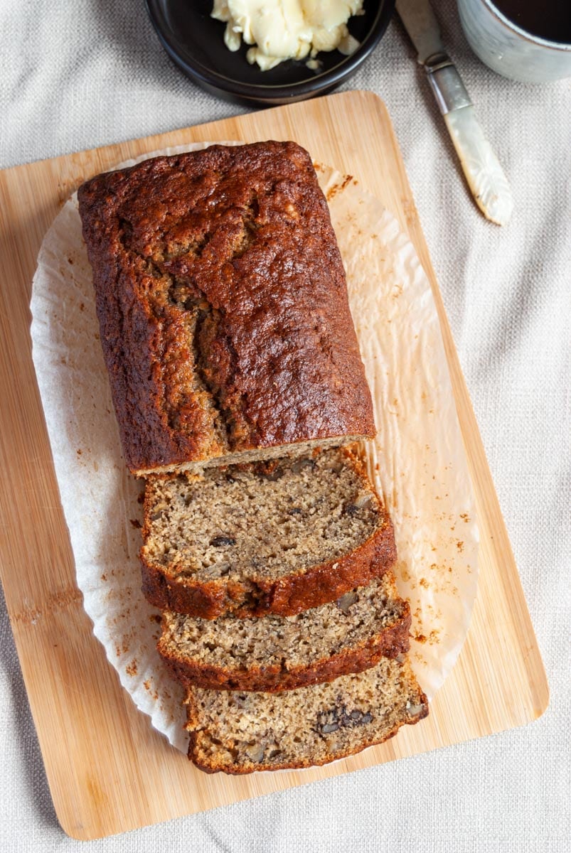 a banana loaf cake on a wooden board, a pot of butter and a light blue cup of coffee on a beige linen napkin.