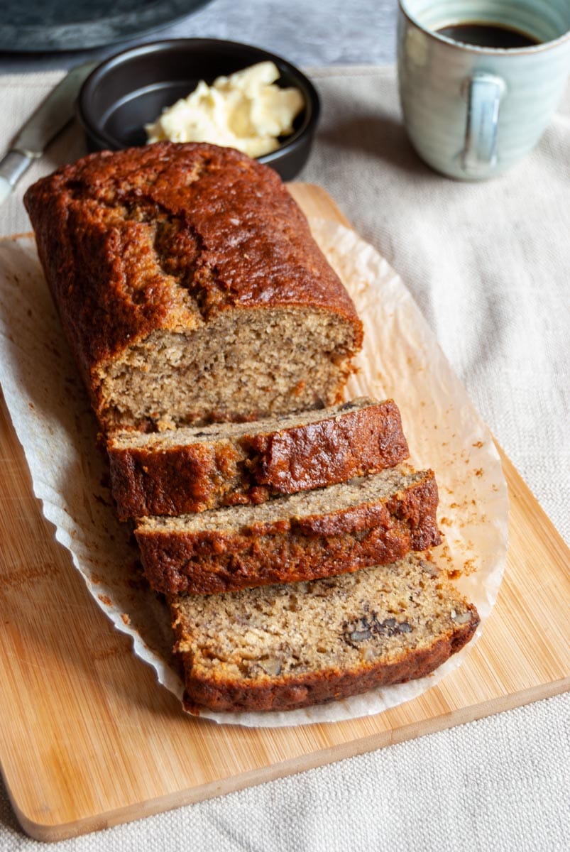 a banana loaf cake partially sliced on a wooden board with a blue cup of coffee and a black bowl of butter with a knife.