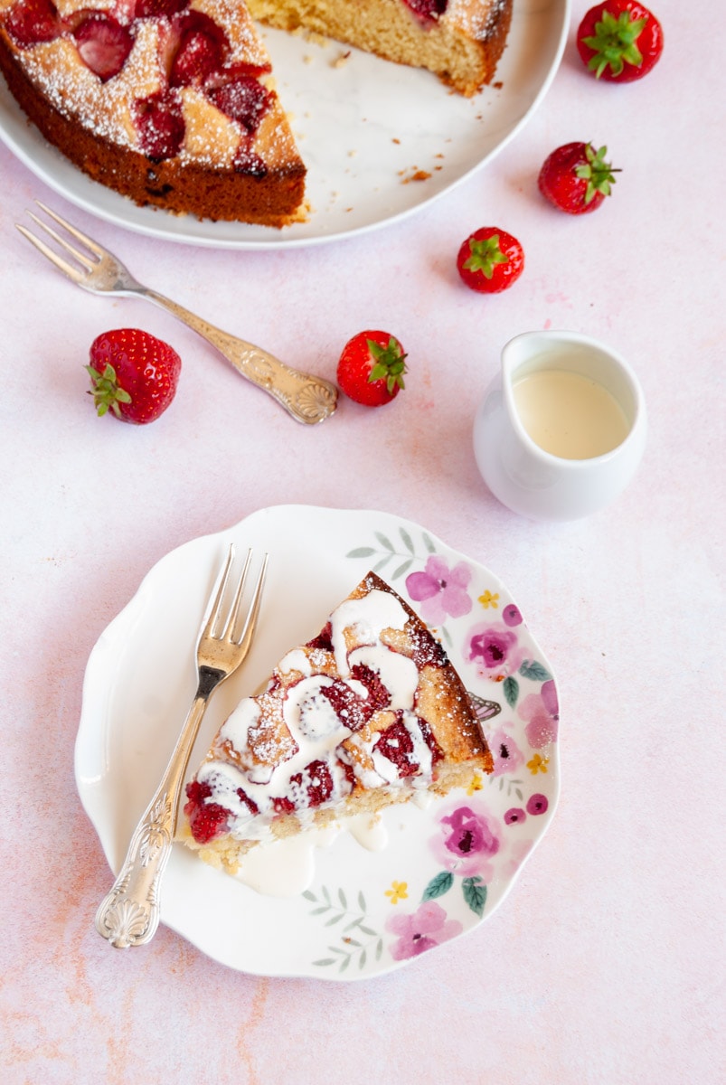 a slice of strawberry cake drizzled with cream on a floral plate with a small white jug of cream.