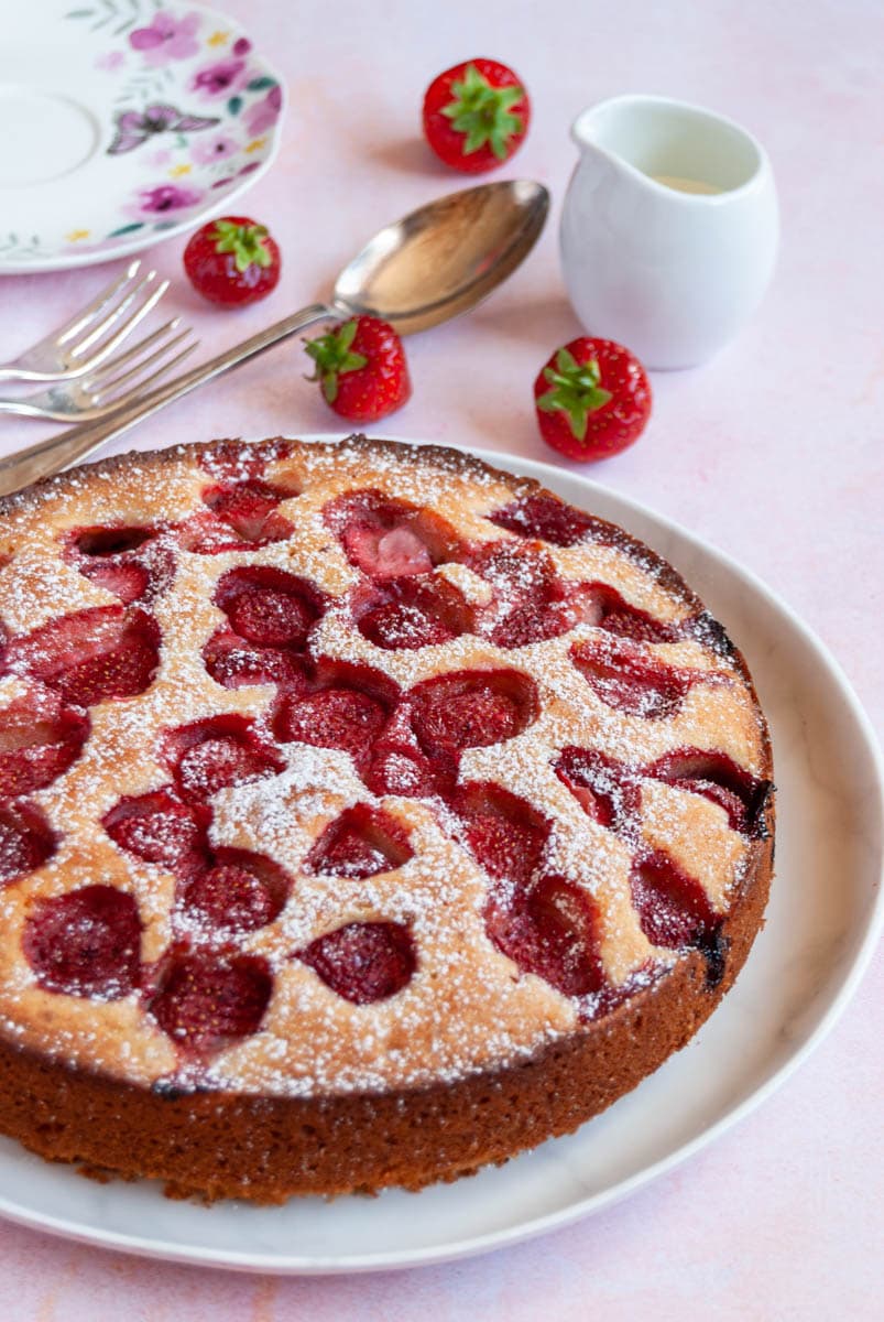 a round strawberry cake on a white plate, a small jug of cream and a floral plate with a spoon.