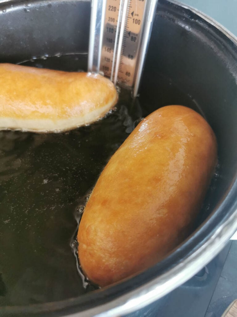 two long doughnuts frying in a large pan with a sugar thermometer
