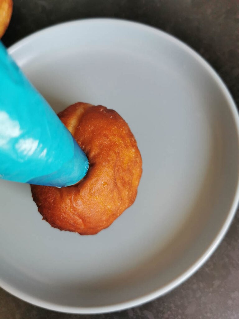 A doughnut on a grey plate being filled with cream using a blue piping bag