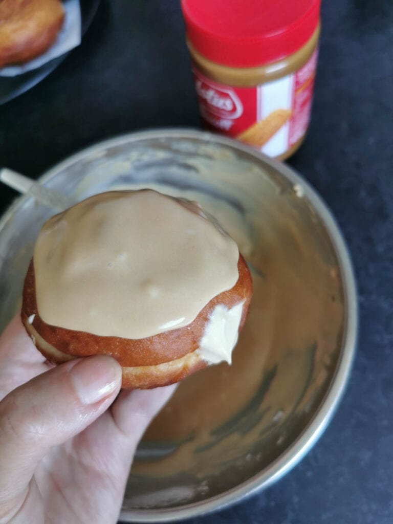 a hand holding a cream filled doughnut topped with Biscoff glaze, a silver bowl of biscoff icing and a jar of Biscoff spread.