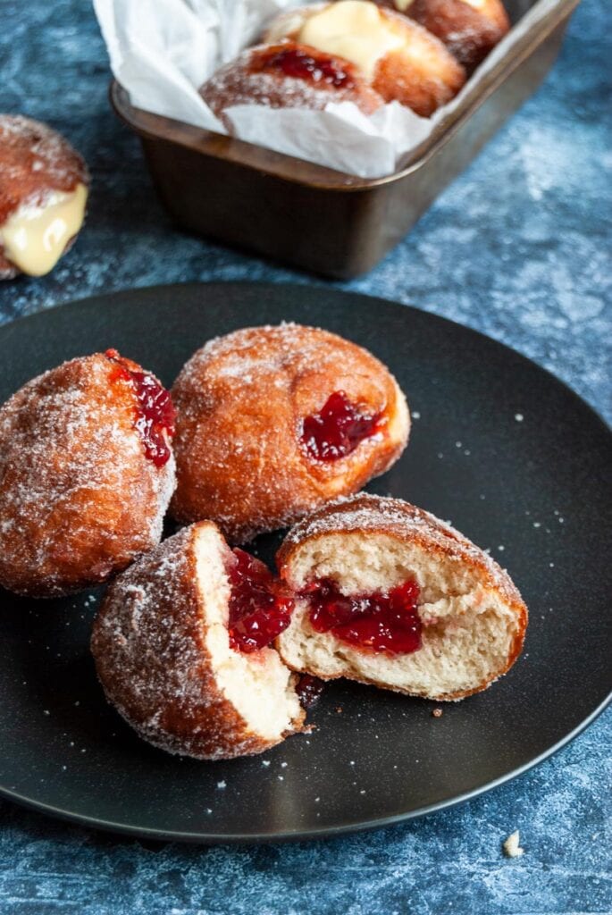 three jam doughnuts on a black plate.  One is cut open to reveal the jam filling.