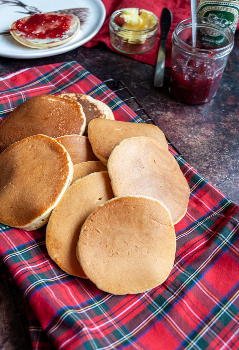 freshly made pancakes on a tartan cloth and wire cooling rack, a jar of raspberry jam, a pot of butter and a pancake spread with butter and jam on a white plate.