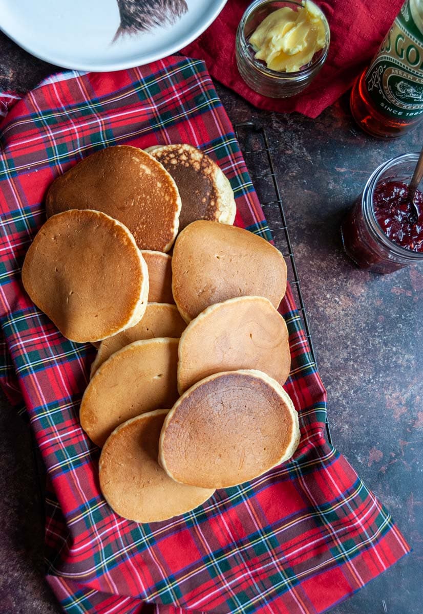 golden pancakes on a tartan tablecloth and wire cooling rack, a jar of raspberry jam, a pot of butter and a bottle of golden syrup.