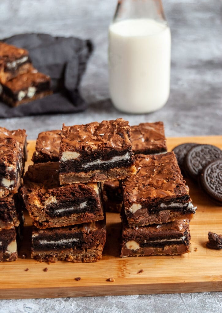 a stack of oreo cookie stuffed cookie dough chocolate brownies on a wooden board with a glass bottle of milk in the background.