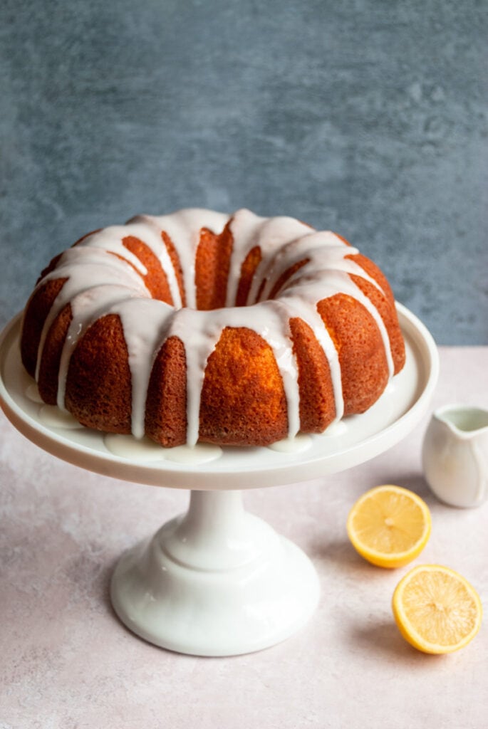 a lemon bundt cake with a lemon icing drizzle on a white cake stand, a lemon sliced in half and a small white jug.