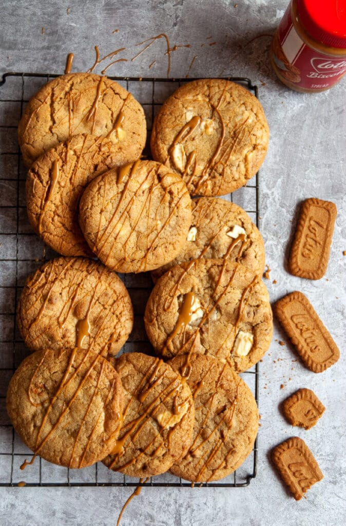 A flat lay photo of a wire rack with Biscoff and white chocolate cookies and a jar of Biscoff on a grey background