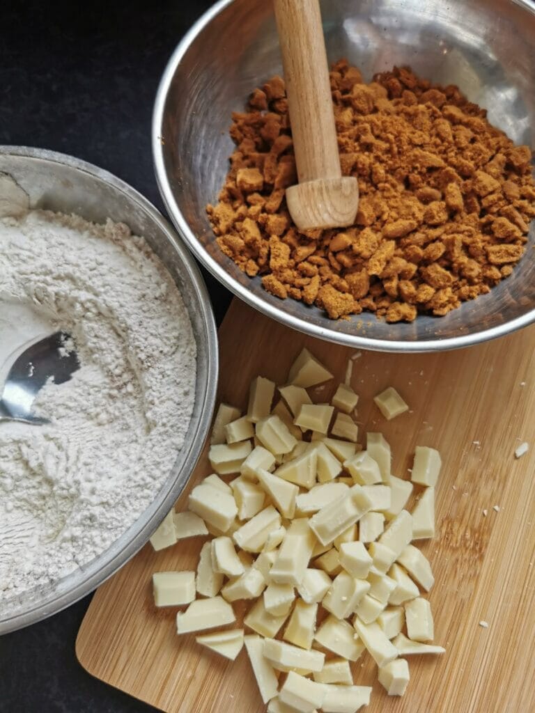 A bowl of flour, a silver bowl of crushed biscoff cookie crumbs and chopped white chocolate on a wooden board