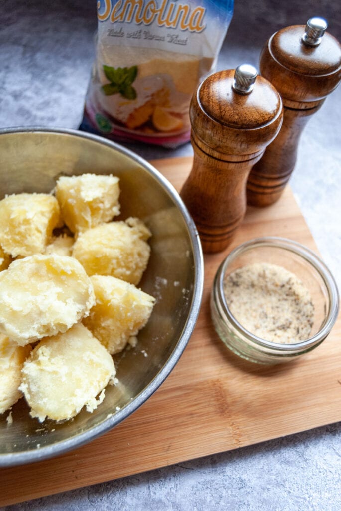 a silver bowl of freshly boiled potatoes for roasting, a pot of roast potato seasoning, a packet of semolina and wooden salt and pepper mills on a wooden chopping board.
