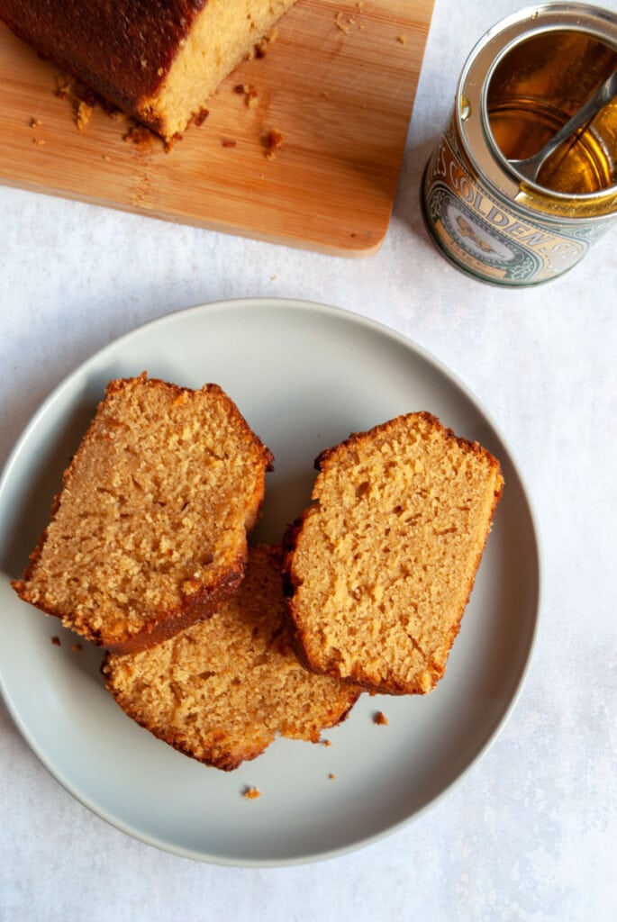 three slices of syrup loaf cake on a blue plate, a partially cut syrup cake on a wooden board and a tin of golden syrup.