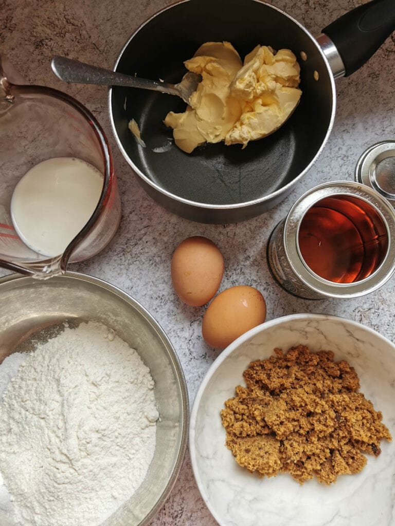 a saucepan of butter, a white bowl of brown sugar, two eggs, a bowl of flour, a tin of golden syrup and a jug of milk.