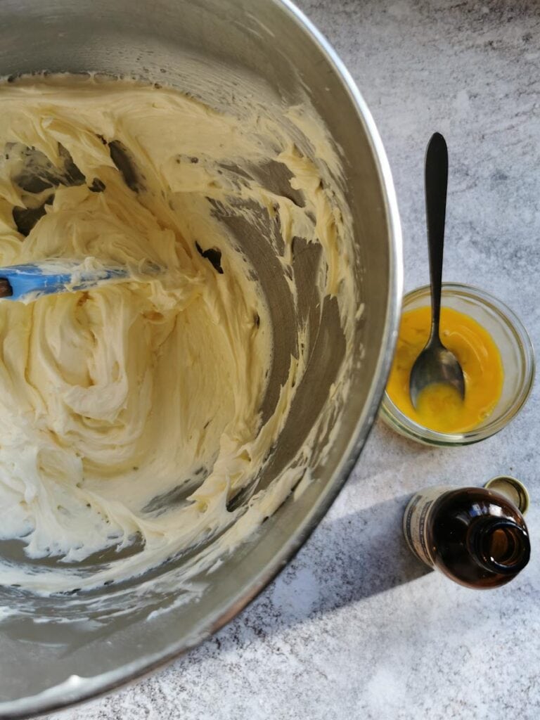A silver bowl of creamed butter and sugar, a small glass pot of beaten egg and a bottle of vanilla extract.