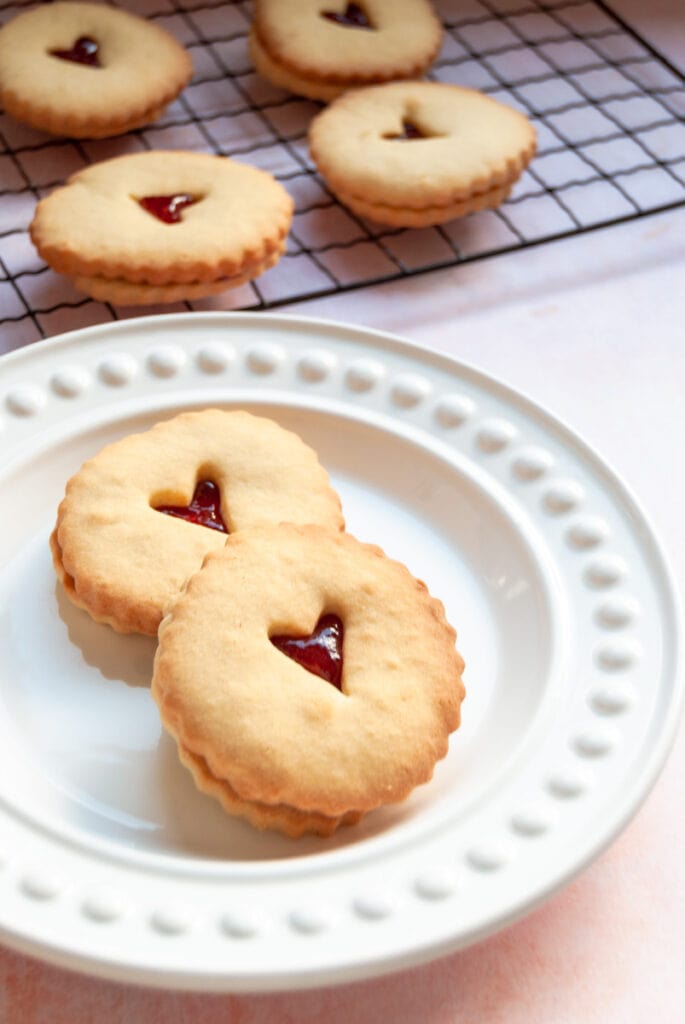 two jam filled biscuits with a heart cut out middle on a white plate and a cooling rack with more biscuits in the background.