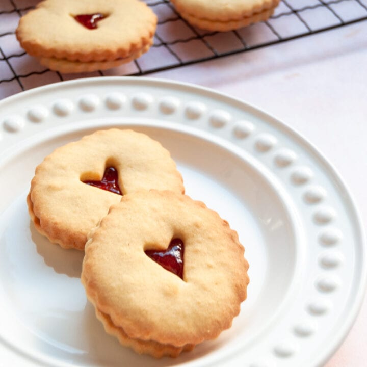 two jam filled biscuits with a heart cut out middle on a white plate and a cooling rack with more biscuits in the background.