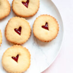 six cookies with a jam filled heart centre on a white plate.