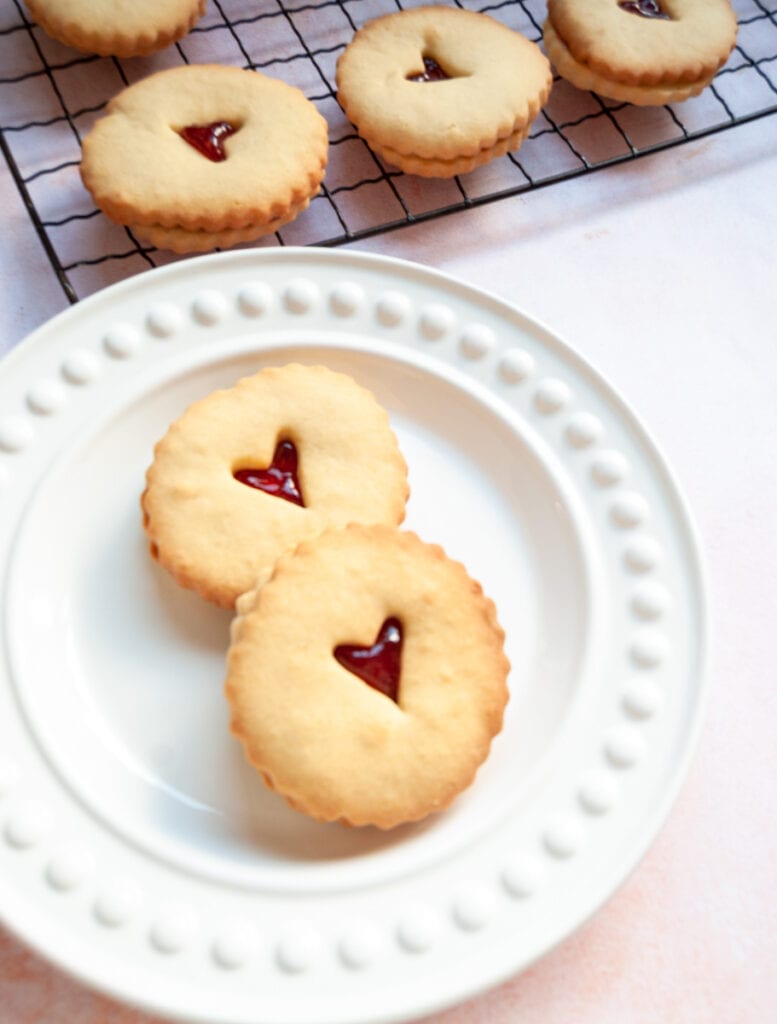 two jammie dodger biscuits on a white plate.  More biscuits on a wire cooling rack are in the background.