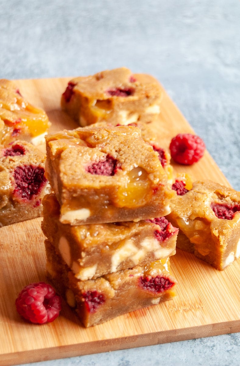 a stack of three lemon blondies with white chocolate chunks, raspberries and lemon curd on a wooden board.