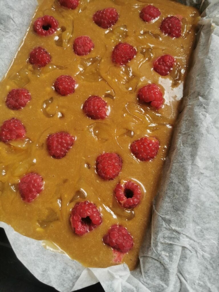 blondie batter with fresh raspberries and lemon curd swirls in a lined baking tin.