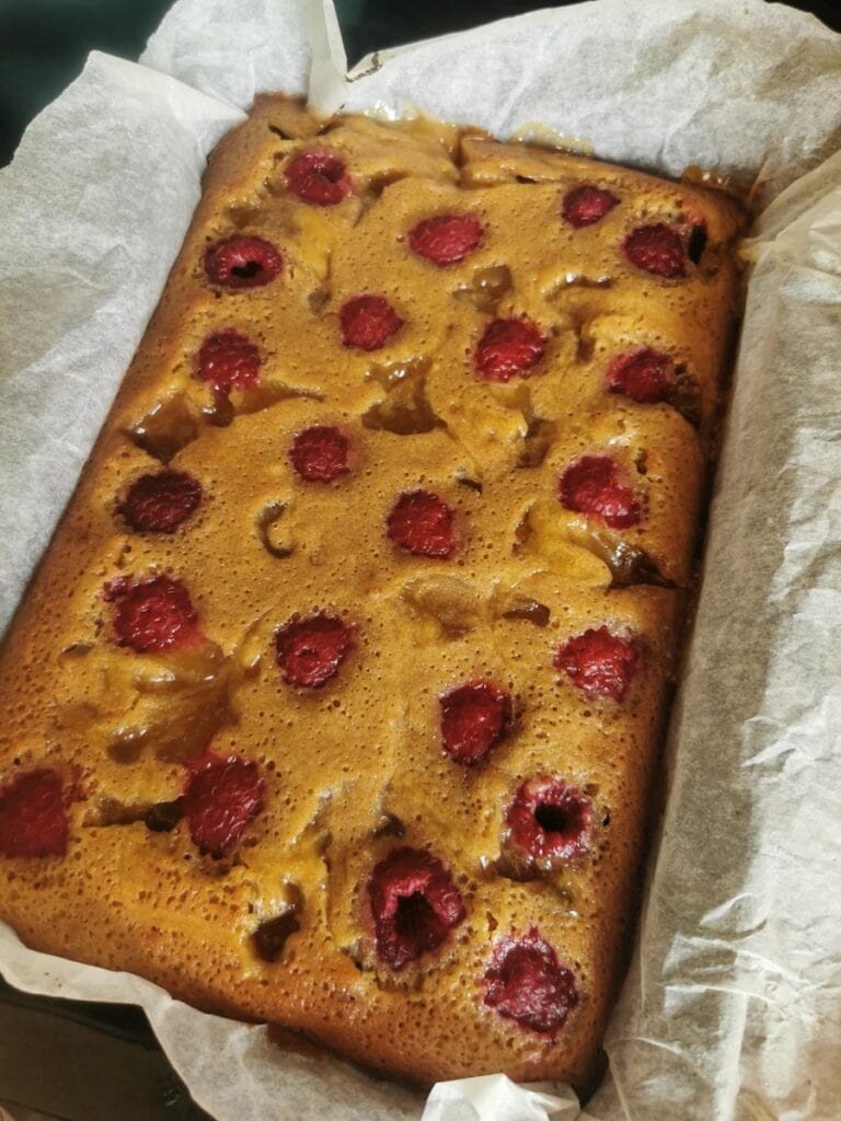 A blondie bar with fresh raspberries and lemon curd swirls in a lined baking tin.