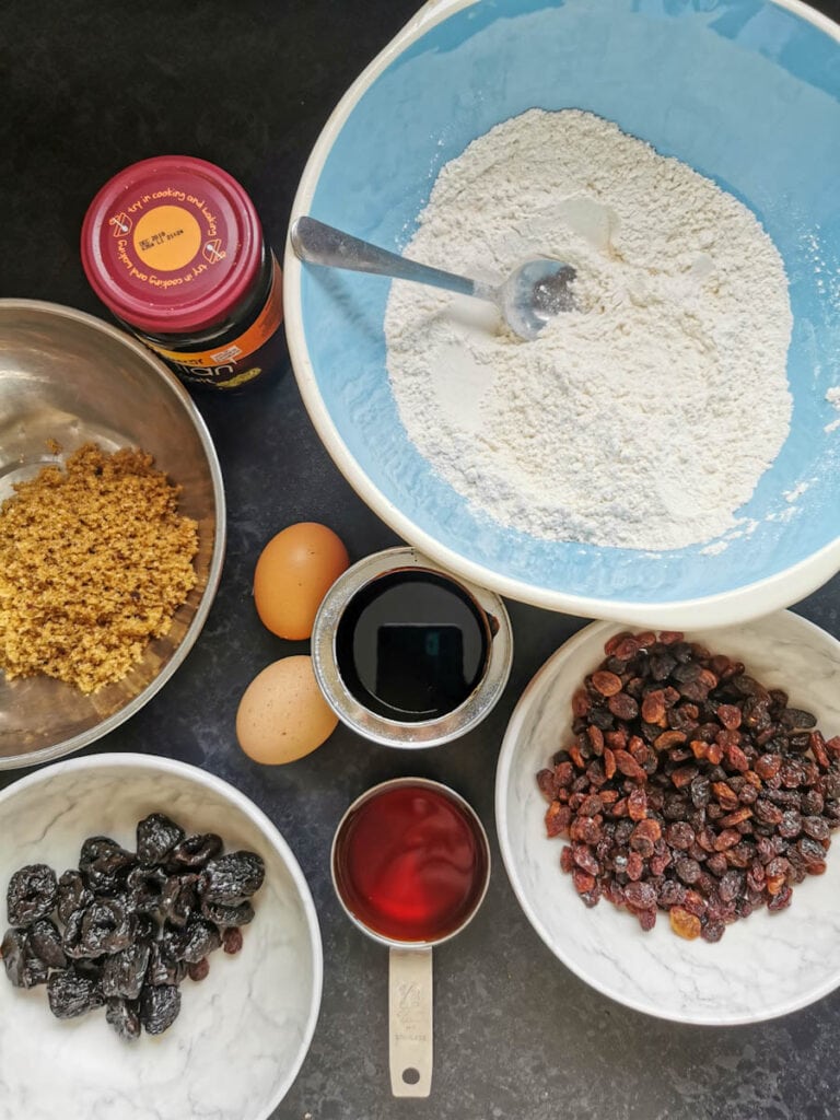 A mixing bowl of flour, bowls of prunes and raisins, two eggs, a tin of treacle, a jar of malt extract and a bowl of brown sugar.