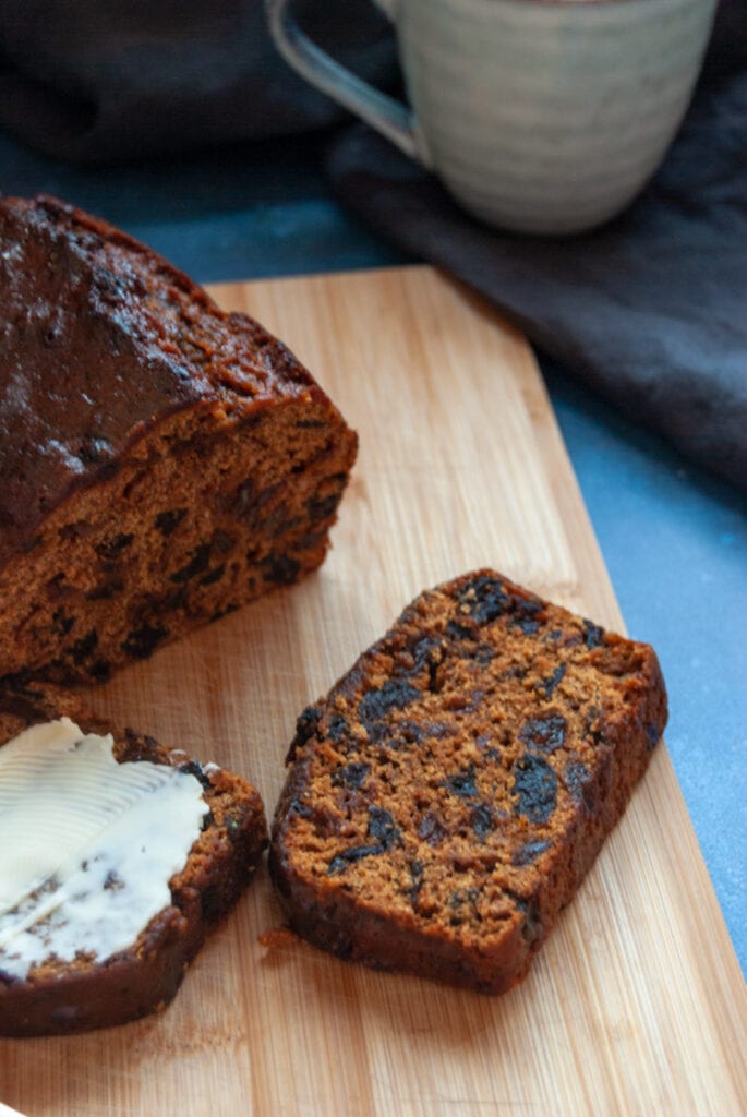a fruit malt loaf with two cut slices - one spread with butter - on a wooden chopping board.