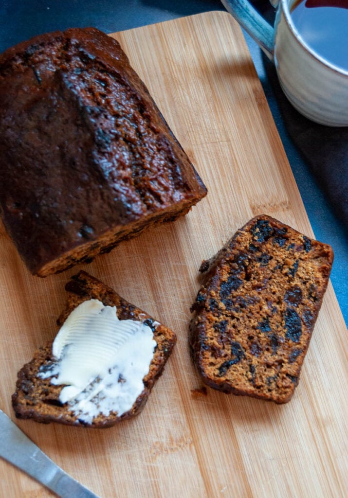 a malt fruit loaf with two cut slices - one spread with butter - on a wooden chopping board.