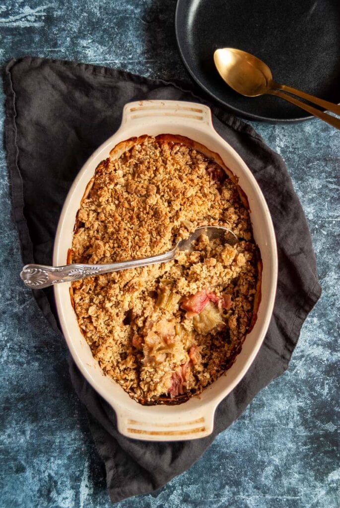 a rhubarb crumble in an oval baking dish with a silver spoon on a grey linen cloth.