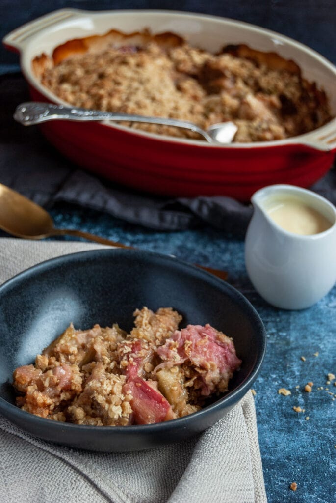 a bowl of rhubarb crumble with an oaty topping, a white jug of custard and an oval red dish of the crumble.