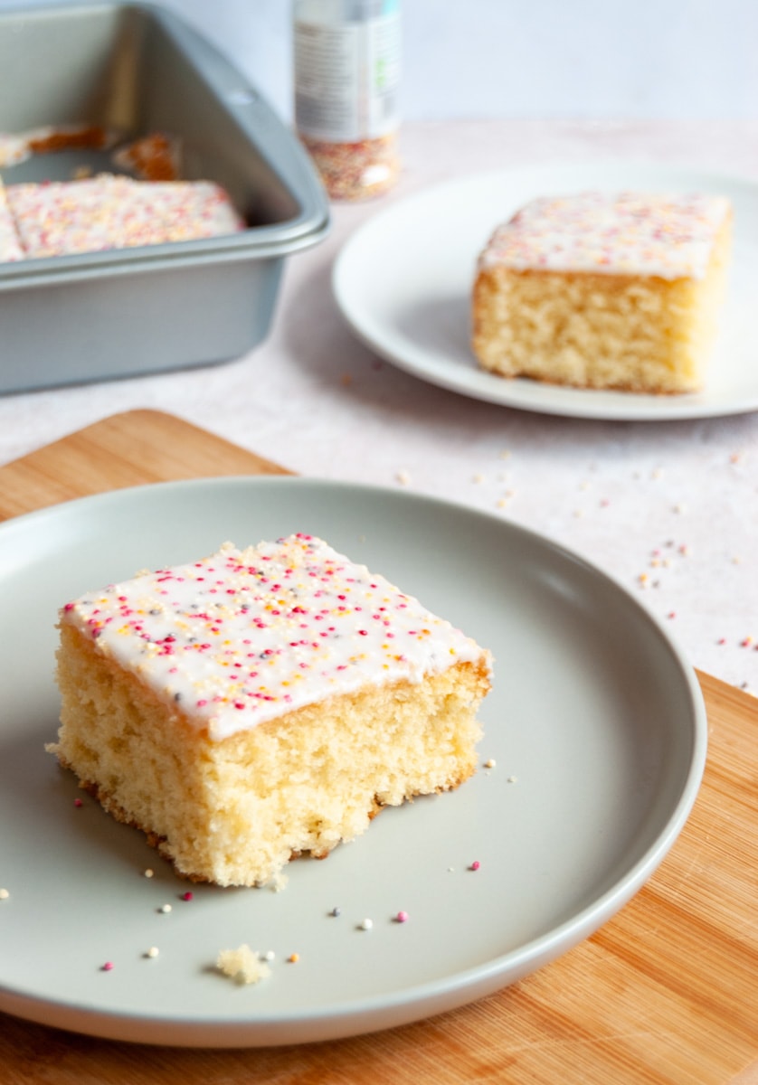 a square of vanilla sponge cake covered in white icing and coloured sprinkles on a light grey plate and wooden board.
