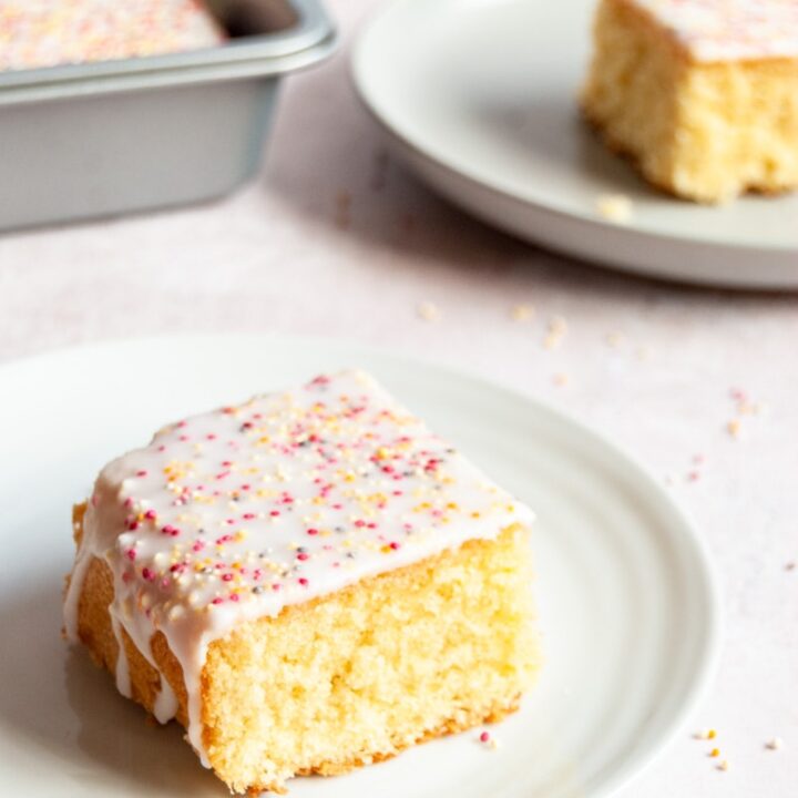 a square piece of vanilla sponge cake with white icing and coloured sprinkles on a white plate.