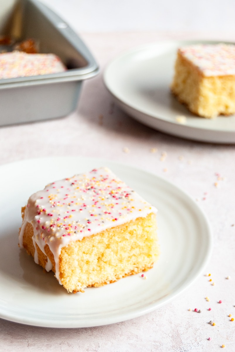 a square piece of vanilla sponge cake with white icing and coloured sprinkles on a white plate.