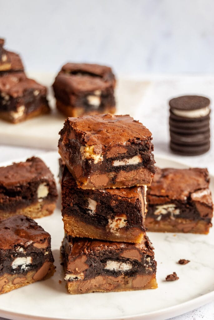 a stack of chocolate chip cookie dough brownies stuffed with Oreo cookies on a white plate.