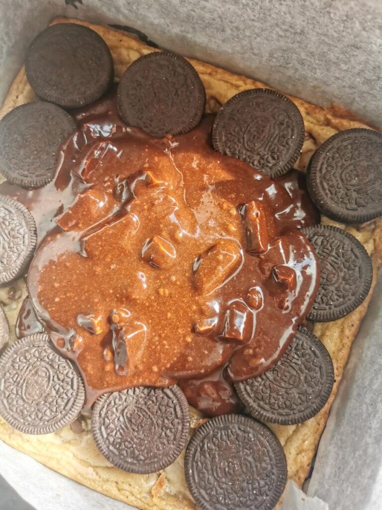 chocolate brownie batter being poured over a layer of oreo cookies and chocolate chip cookie dough.