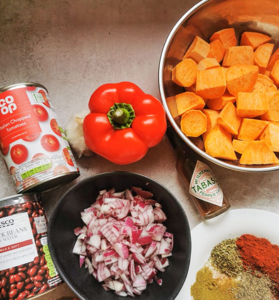 bowls of chopped sweet potato, chopped red onion, a red pepper, tinned tomatoes and black beans,a bottle of Tabasco sauce and a plate of spices.