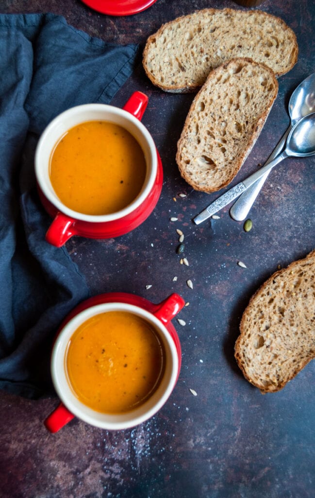 two red bowls of sweet potato soup, a black napkin, two silver spoons and two slices of brown bread.