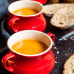 two red bowls filled with sweet potato soup, slices of bread and a salt and pepper mill