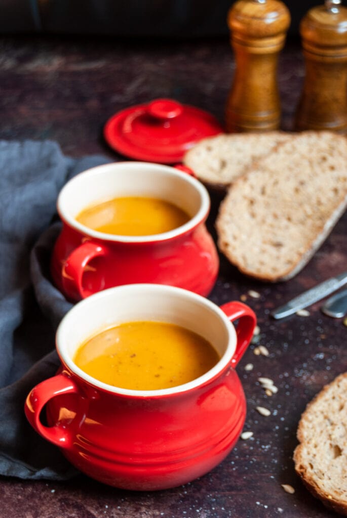 two red bowls of sweet potato and red pepper soup, crusty brown bread, two spoons and a wooden salt and pepper mill.