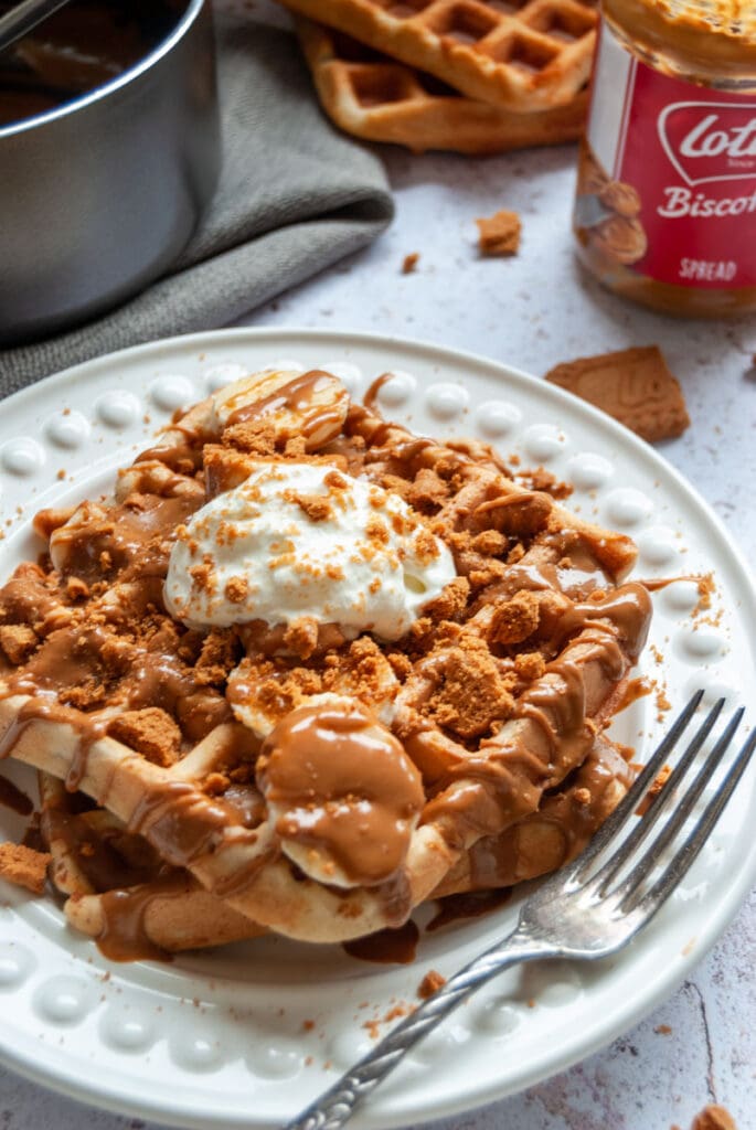 two buttermilk waffles on a white place topped with sliced bananas, melted biscoff spread and whipped cream.