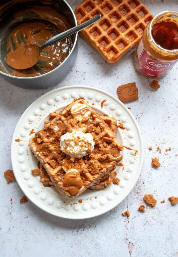 two buttermilk waffles on a white plate with sliced bananas, melted Biscoff spread and whipped cream.