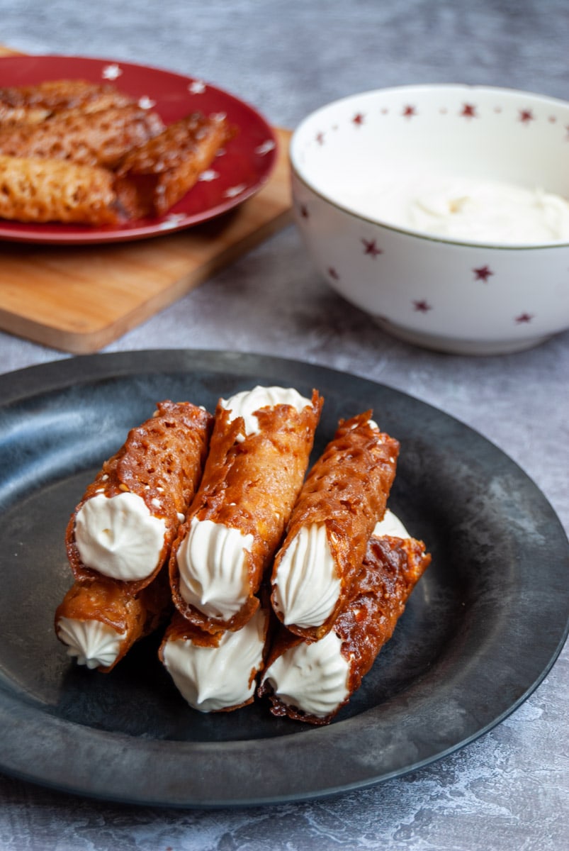 brandy snap biscuits filled with whipped cream on a black plate, a white bowl of whipped cream and a red plate with more brandy snaps.