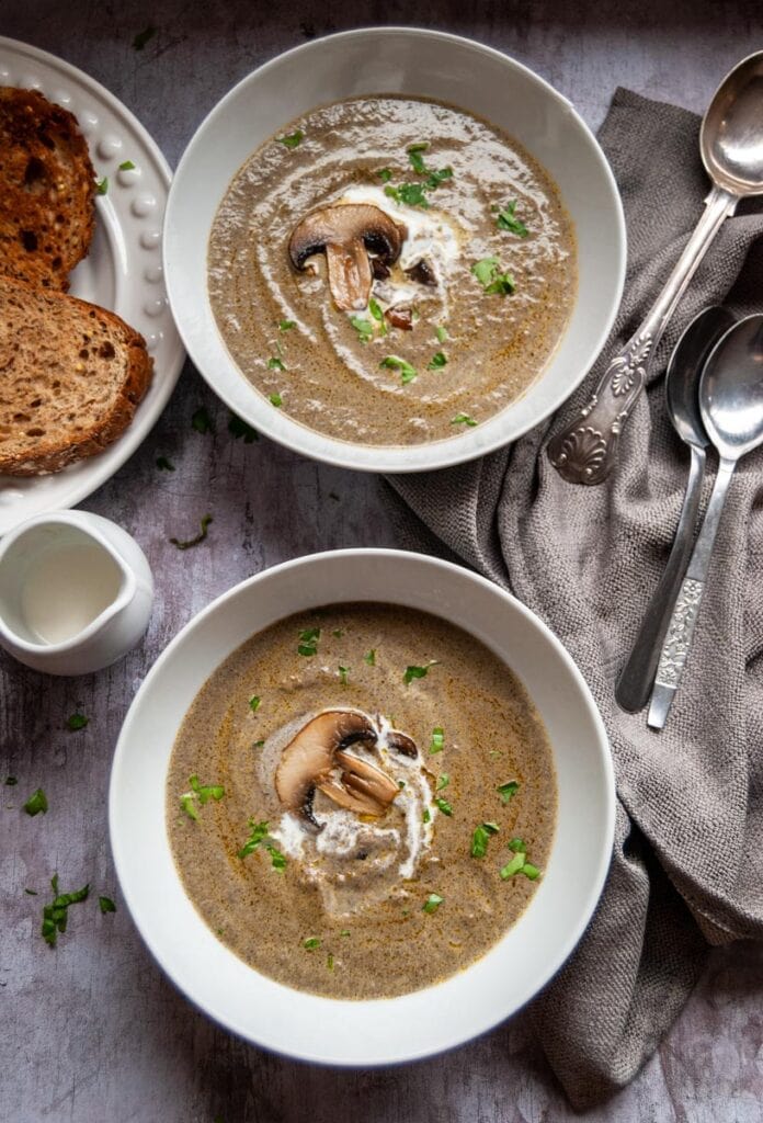 two white bowls of mushroom soup, a white jug of cream, two silver spoons and a white plate of brown bread.