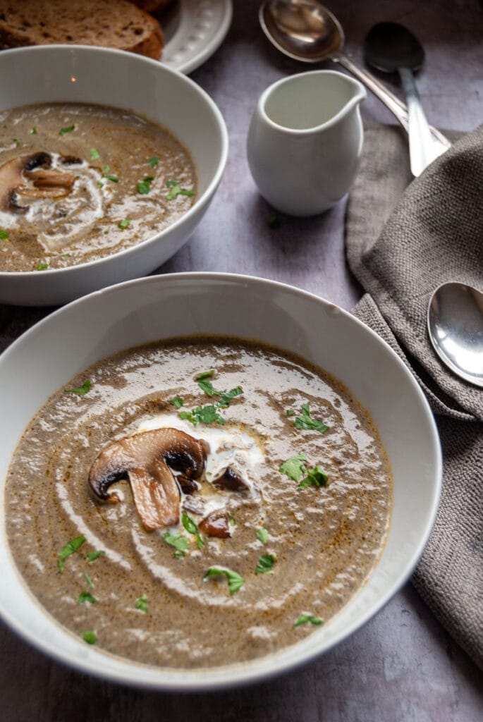 two white bowls of mushroom soup topped with sliced mushrooms and a swirl of cream, a small white pot of cream and a beige napkin with silver spoons.