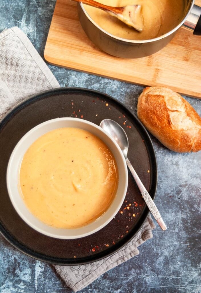 a bowl on parsnip soup on a black plate with a silver spoon, a crusty bread roll and a small pan of soup with a wooden spoon.