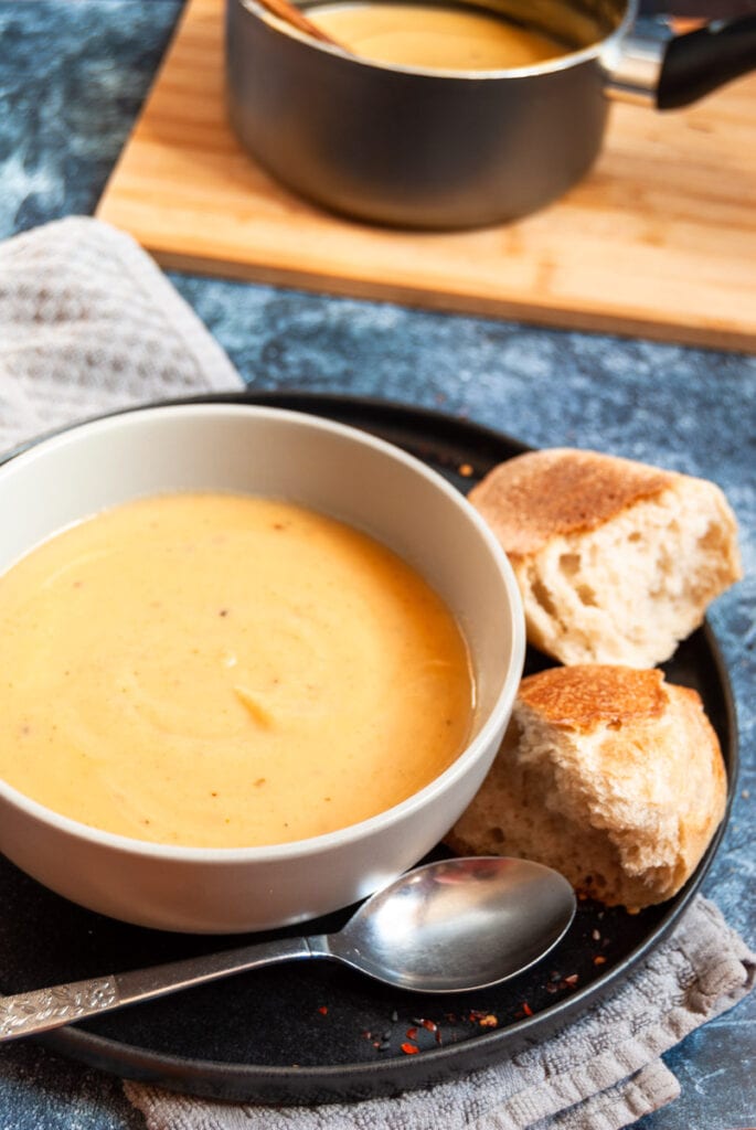 a bowl of parsnip soup on a black plate with crusty bread and a silver spoon. A pot of soup with a wooden spoon on a wooden board in the background,